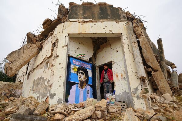 Сирийский художник Азиз Асмар стоит у рисунка на стене разрушенного дома знаменитого аргентинского футболиста Марадоны (26 ноября 2020). Бинниш - Sputnik Армения
