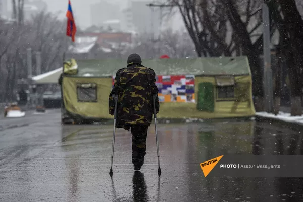 Ветеран войны с ампутированной ногой на фоне палаток, установленных на заблокированном оппозиционерами проспекте Баграмяна (1 марта 2021). Еревaн
