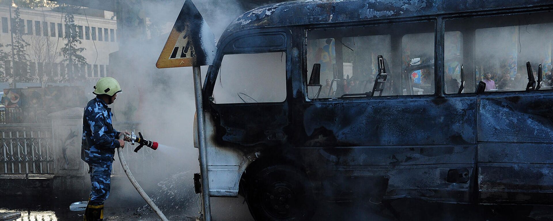 Сирийский пожарный тушит сгоревший автобус на месте взрыва (20 октября 2021). Дамаск - Sputnik Армения, 1920, 20.10.2021