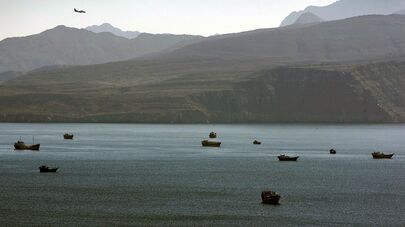 Самолет пролетает над горами к югу от Ормузского пролива, в то время как торговые корабли пришвартованы в водах Персидского залива недалеко от города Хасаб в Омане