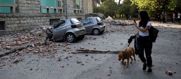 Женщина с собакой проходит мимо разрушенных автомобилей и поврежденного здания после землетрясения (21 сентября 2019). Тиранa - Sputnik Արմենիա