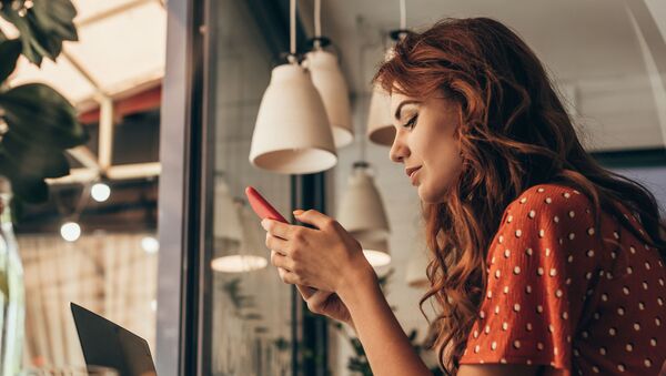 Рыжеволосая девушка в красном платье сидит в кафе и смотрит на свой смартфон - Sputnik Армения