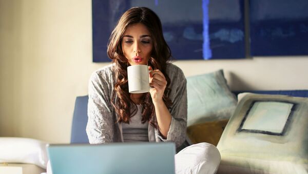 Девушка брюнетка с чашкой чая сидит на кровати и смотрит видео по ноутбуку - Sputnik Армения